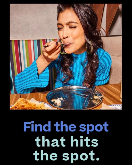 Woman with dark hair in a blue top enjoys a dessert with a spoon. Table holds a metal plate and fork. Backdrop has striped fabric.