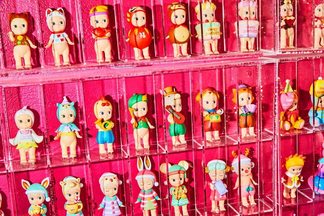 Rows of Sonny Angel collectible dolls with varied headwear fill the frame against a vibrant pink background, each unique and colorful.