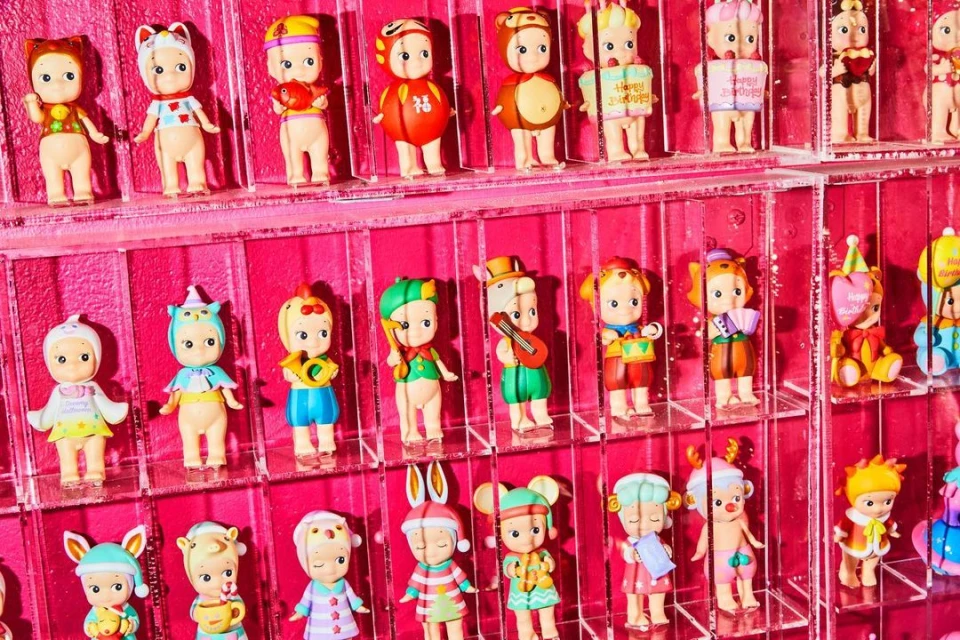 Rows of Sonny Angel collectible dolls with varied headwear fill the frame against a vibrant pink background, each unique and colorful.