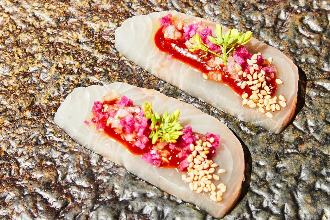 Overhead shot featuring two slices of fish with red sauce, pink relish, and green sprouts, resting on a stone surface.