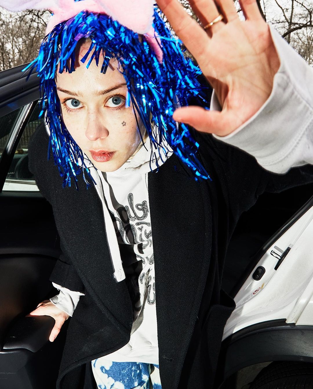 Young woman poses in blue tinsel wig, pink bunny ears, and black coat with car interior in background. Hand is up in the foreground.