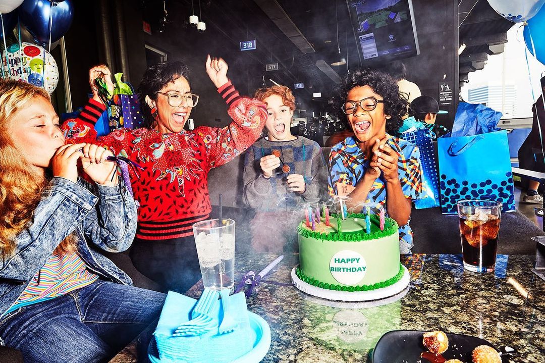 A group of children and an adult celebrate a birthday with a green frosted cake, balloons, and gifts in an indoor party setting.