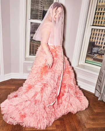 Woman in a pink gown and veil poses indoors by windows overlooking a cityscape, creating an ethereal, fashion-forward bridal style.