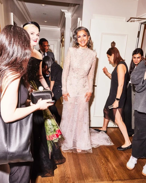 Elegant older woman in long embellished gown poses at an event, surrounded by a group of people in an indoor setting.