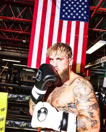 Man wearing boxing gloves stands in a boxing ring under a US flag. He has tattoos on his arms and is facing forward in a portrait shot.