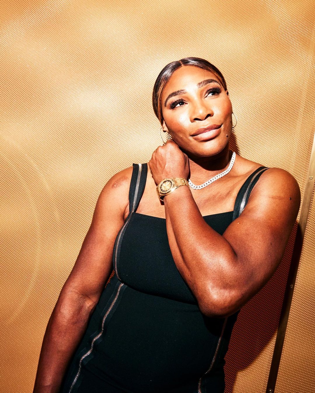 Portrait of Serena Williams in a black dress, gold watch and necklace, posing indoors against a textured gold background.