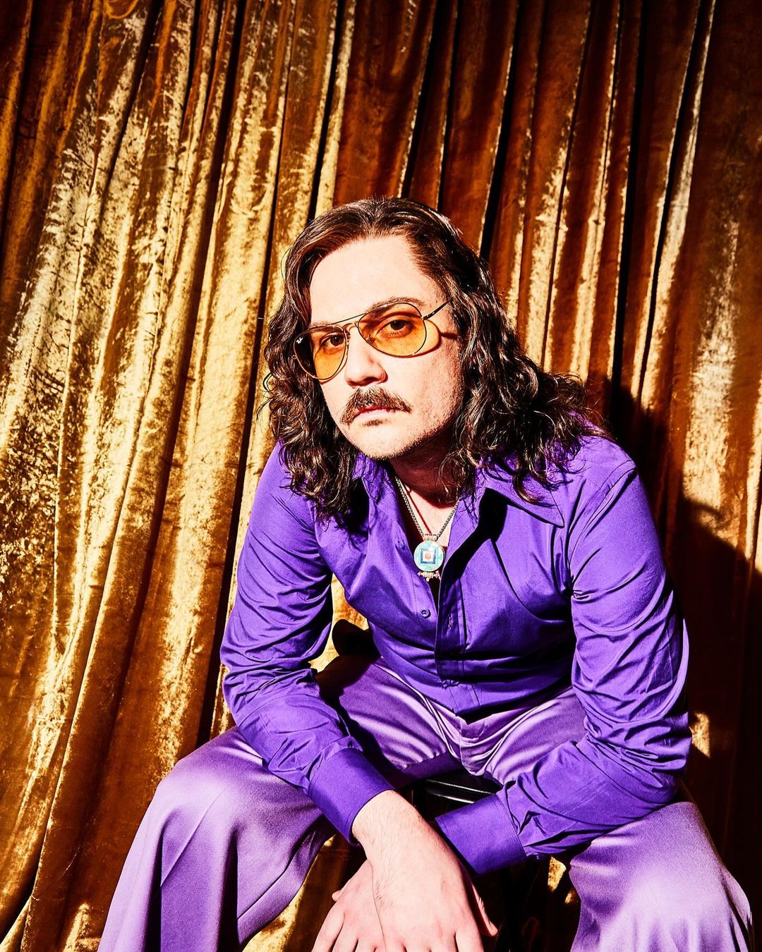 Portrait of a seated adult male wearing a purple satin shirt and pants with tinted glasses, in front of a gold curtain backdrop.
