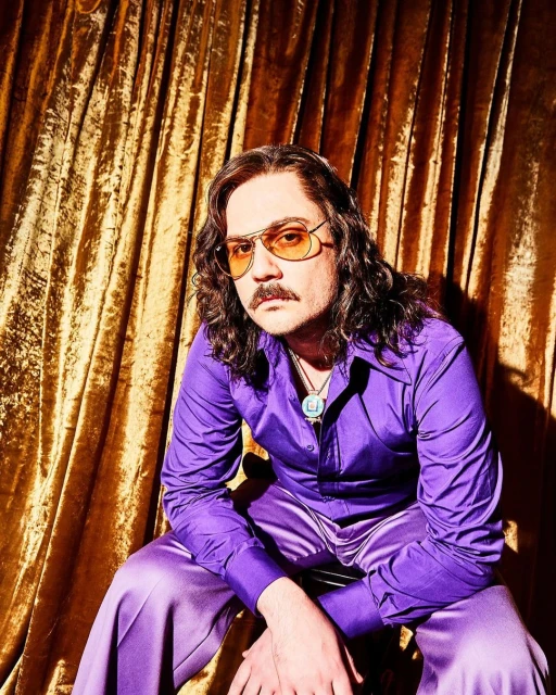 Portrait of a seated adult male wearing a purple satin shirt and pants with tinted glasses, in front of a gold curtain backdrop.