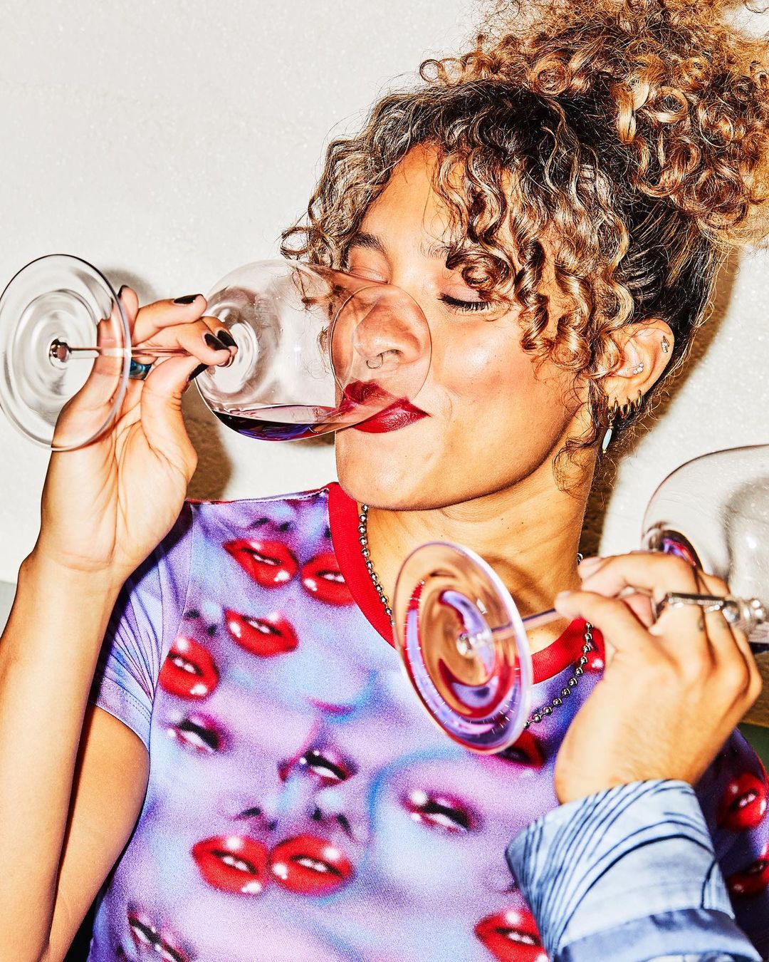 Close-up of a stylish woman with curly hair drinking red wine from a glass, wearing a colorful shirt with lip prints, indoors.