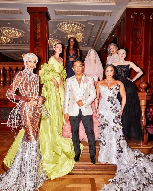 Group of fashion models and a designer pose in elegant gowns on ornate stairs with decorative chandeliers and luxurious architectural details.