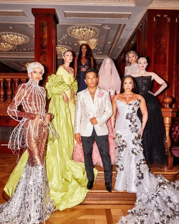 Group of fashion models and a designer pose in elegant gowns on ornate stairs with decorative chandeliers and luxurious architectural details.