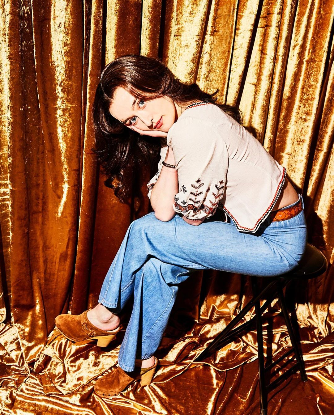 Young woman with long brown hair sits on a stool in front of a gold crushed velvet backdrop, wearing a patterned blouse and blue jeans.