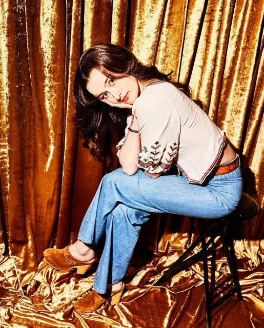 Young woman with long brown hair sits on a stool in front of a gold crushed velvet backdrop, wearing a patterned blouse and blue jeans.