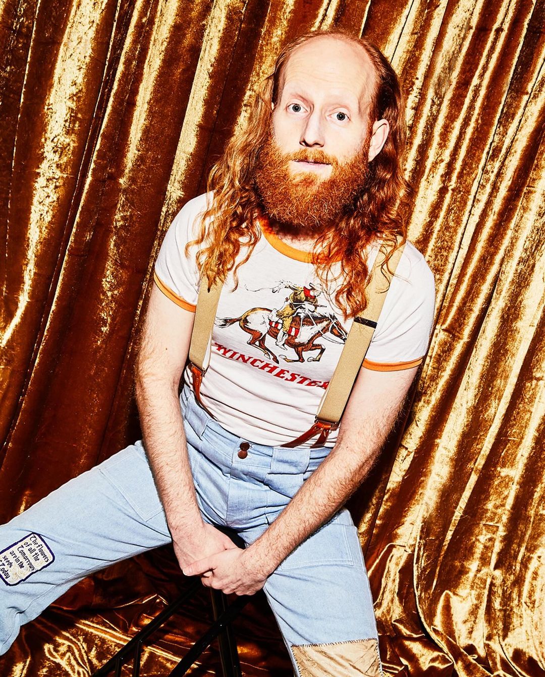 Portrait of a redhead man with a beard in blue jeans and suspenders against a golden curtain backdrop in a studio setting.