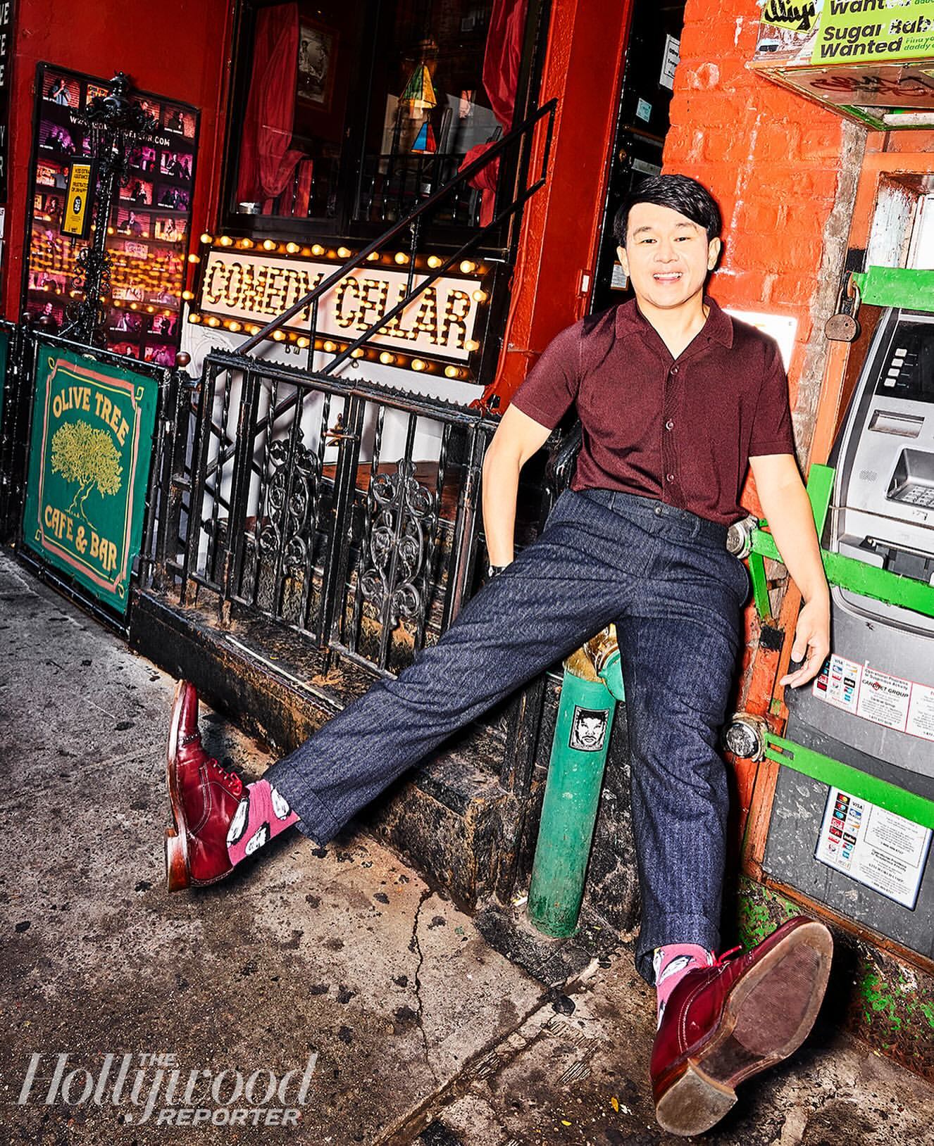 Man leans against an ATM outside Comedy Cellar and Olive Tree Cafe & Bar in the city, wearing red shoes and patterned socks in a portrait.