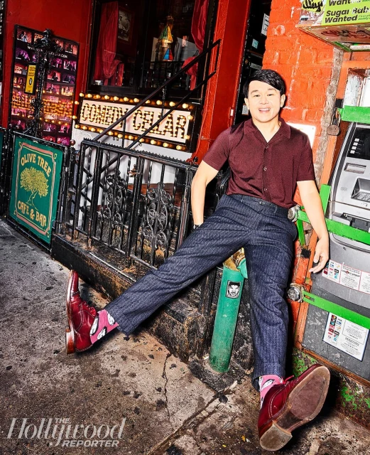 Man leans against an ATM outside Comedy Cellar and Olive Tree Cafe & Bar in the city, wearing red shoes and patterned socks in a portrait.