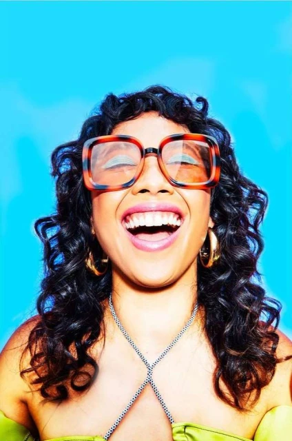 Close-up of a laughing woman with curly dark hair, wearing glasses and a green top against a bright blue background.