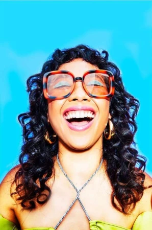 Close-up of a laughing woman with curly dark hair, wearing glasses and a green top against a bright blue background.