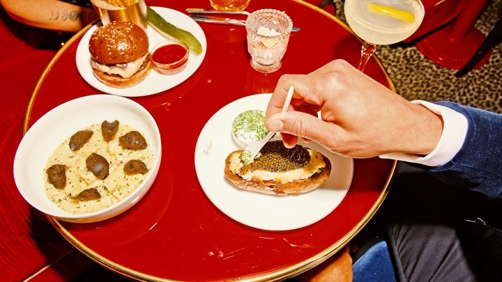 Overhead view of a red table with a burger, truffle pasta, caviar, and a cocktail, presented as gourmet food and beverage options.