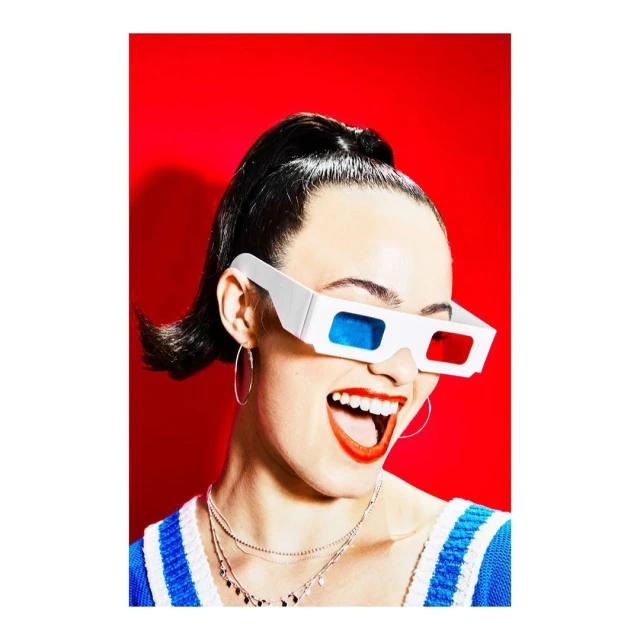 Smiling woman wears 3D glasses against a red background. She has hoop earrings, red lipstick, and a blue and white top.