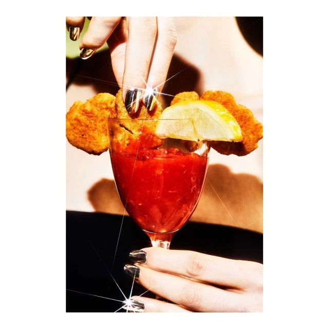 Overhead shot of a hand placing a fried chicken nugget into a stemmed glass of red sauce, garnished with a lemon wedge.