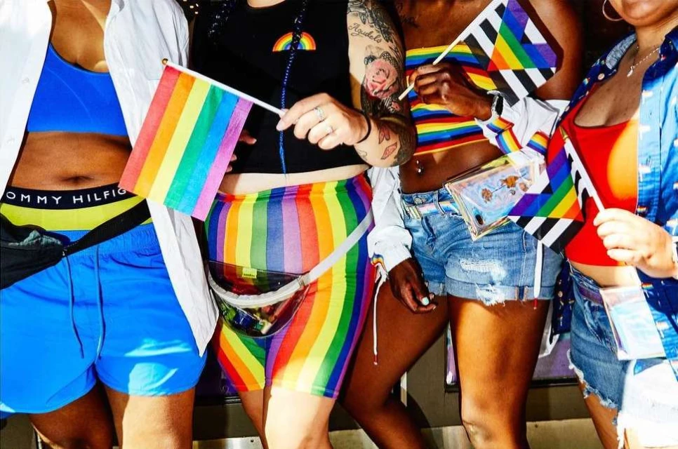 Group of four people wearing rainbow-themed clothing, holding rainbow flags and standing in front of a metallic structure. 