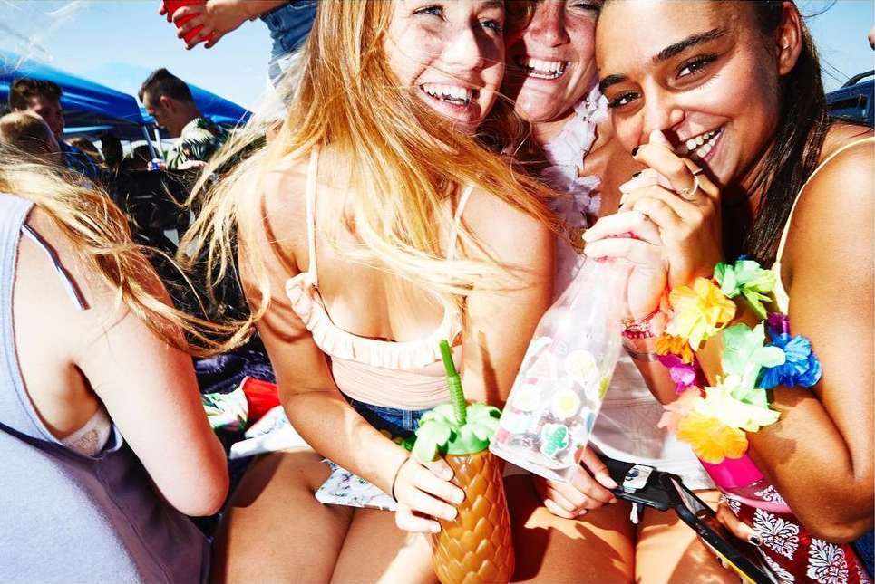 Three smiling women pose together outdoors. Two women wear floral leis. A woman in front holds a pineapple-shaped drink container.