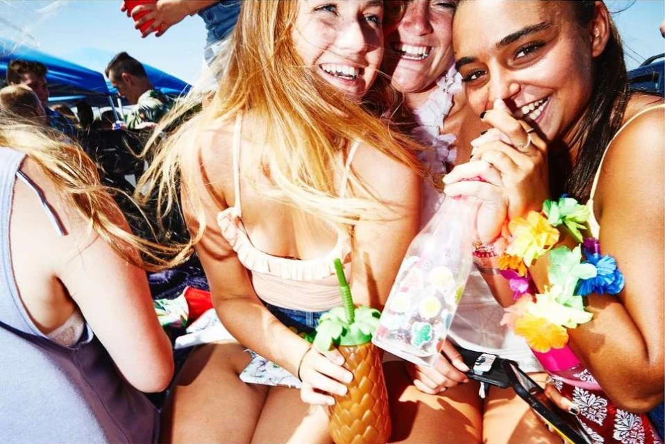 Three smiling women pose together outdoors. Two women wear floral leis. A woman in front holds a pineapple-shaped drink container.