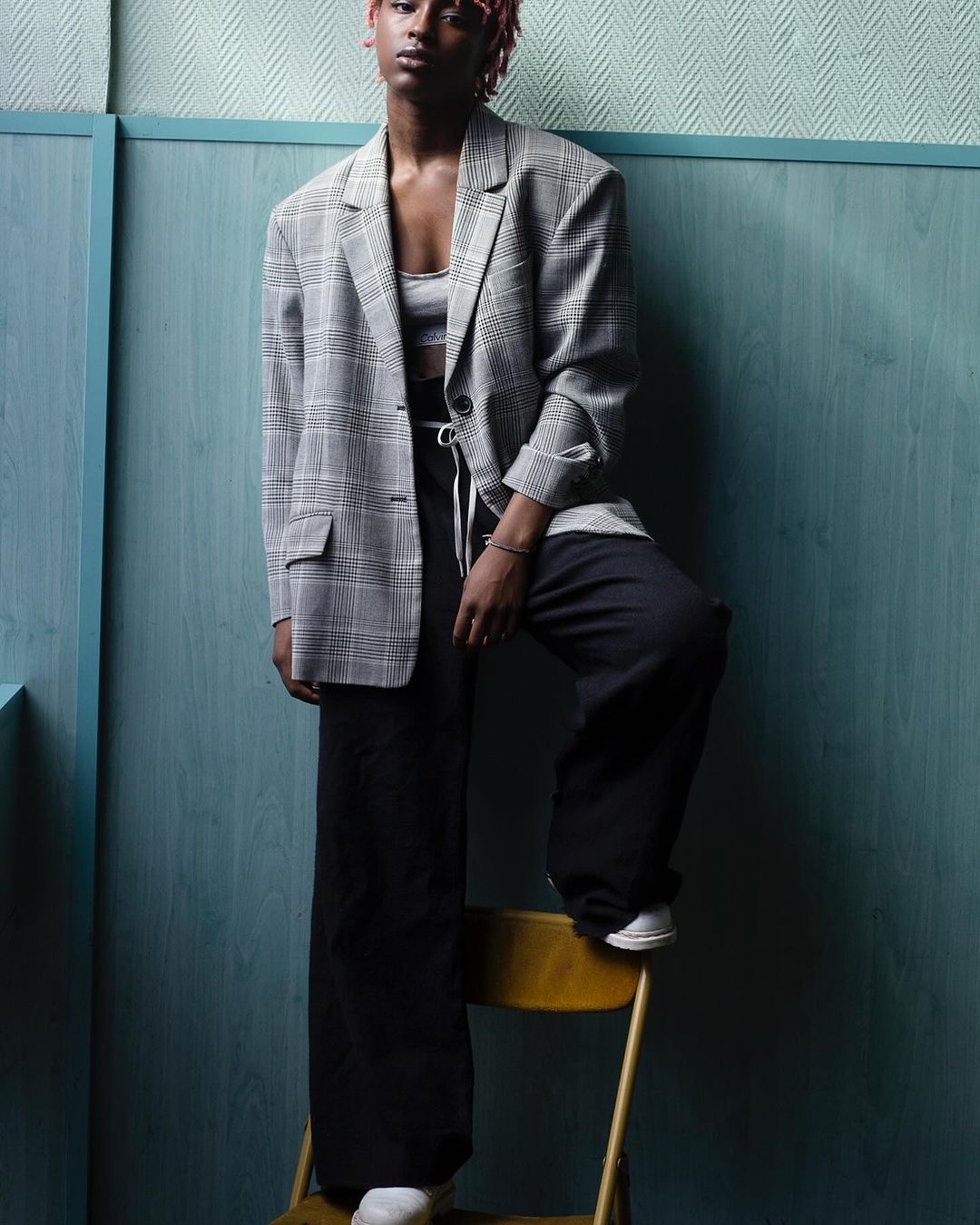 Woman in oversized plaid blazer and black pants poses on a yellow chair in front of a blue paneled wall in a fashion studio shot.