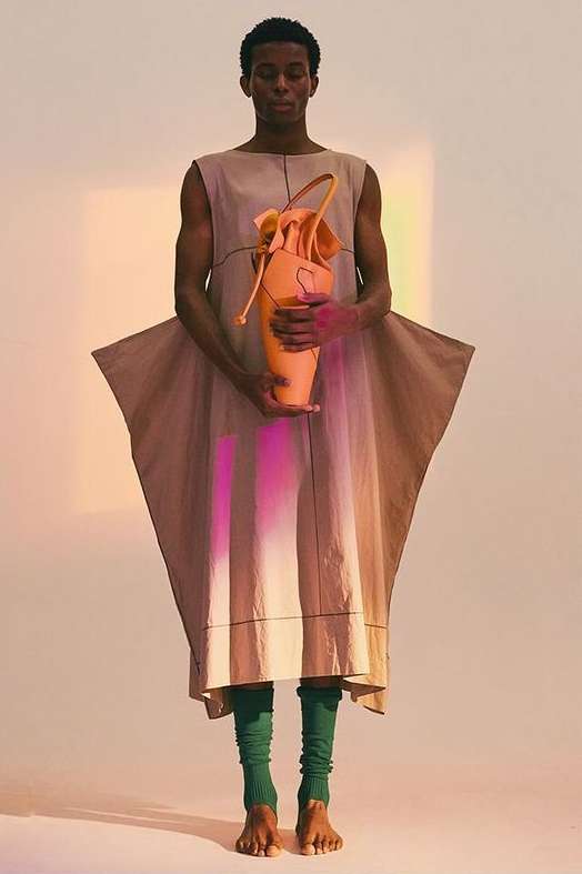 An androgynous person poses in a draped beige gown, holding an orange vase-like bag in a light studio with colorful light streaks.