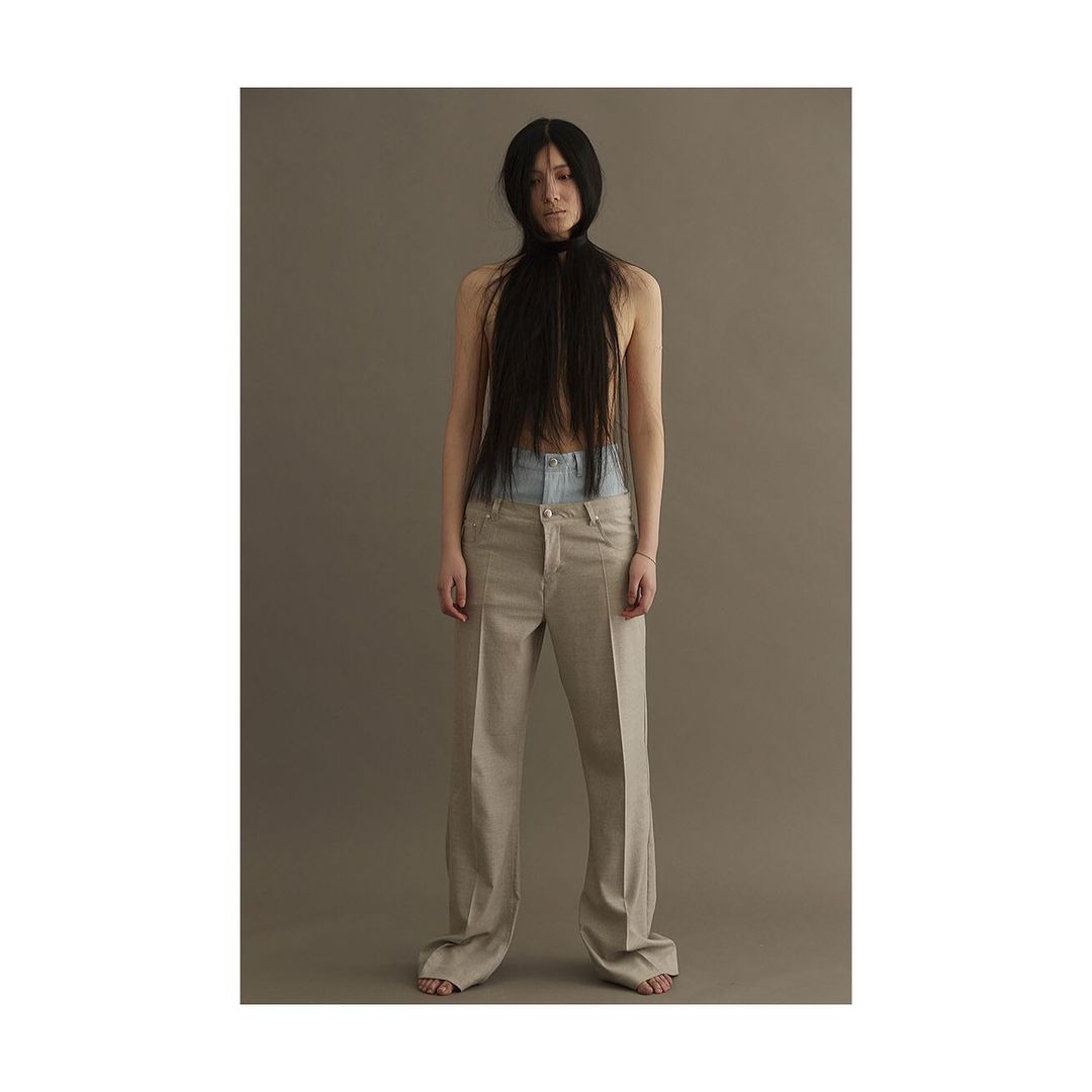 An adult female stands in a photo studio with long black hair covering her face, wearing light blue jeans and beige pants.