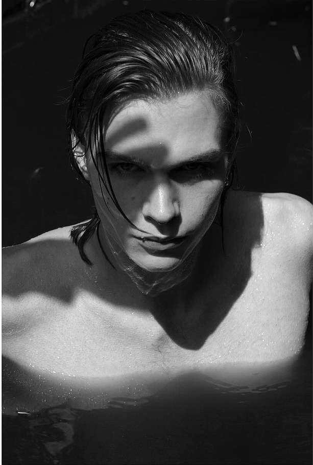 Black and white portrait of a young adult male with wet hair in water, looking directly at the camera with sunlight casting shadows.