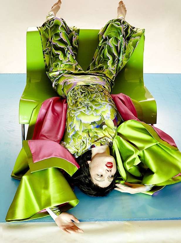 Woman in a green floral outfit with pink accents poses upside down on a bright green chair against a gradient blue backdrop.