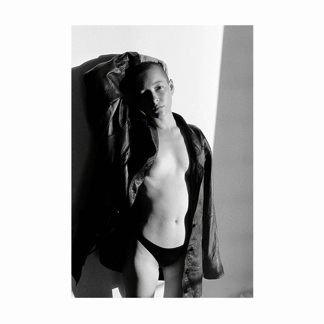 Monochrome portrait of a bald person wearing a leather jacket and black underwear, standing against a bright white wall with shadows.