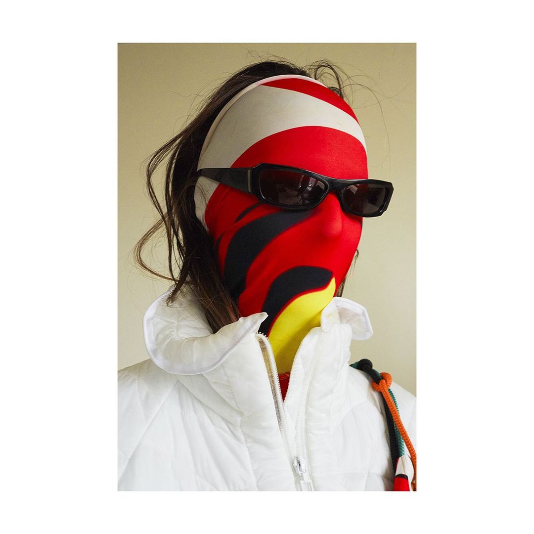 Person wearing black sunglasses and a white puffer jacket, face covered with a red, white, yellow, and black patterned full-face mask.
