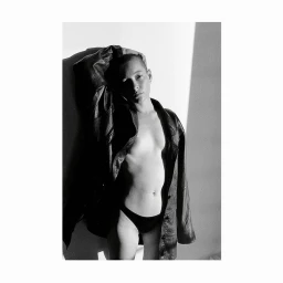 Black and white portrait of a bald woman wearing an oversized leather jacket and underwear posing against a wall