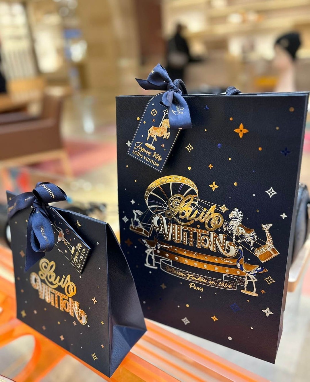 Two Louis Vuitton shopping bags, one larger than the other, are on a blurred surface with gold lettering and a navy-blue background.