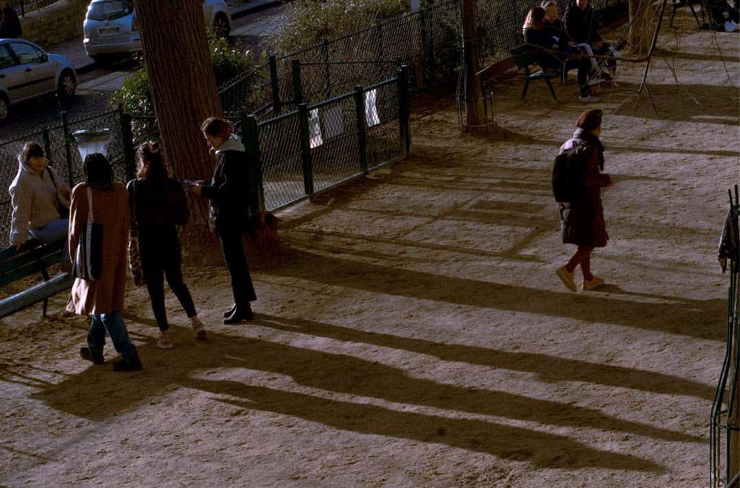 People gathered in an urban park with trees and a metal fence; some sit while others walk, creating shadows on the ground.