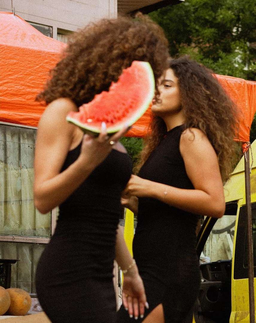 Two women in black dresses outdoors eating watermelon, showcasing a casual, summery vibe with a trailer in the background.
