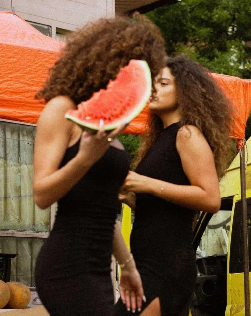 Two women in black dresses outdoors eating watermelon, showcasing a casual, summery vibe with a trailer in the background.