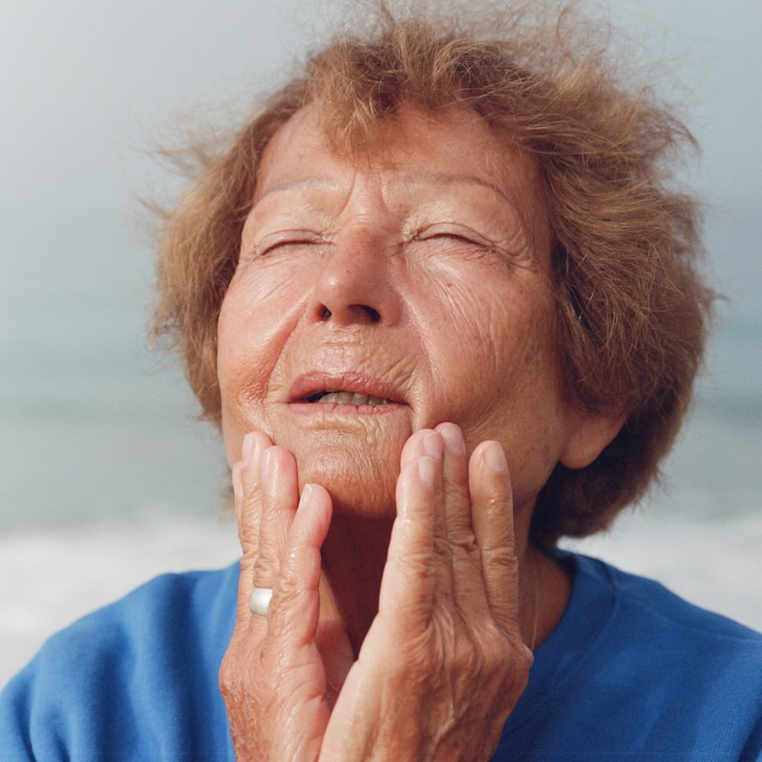 Close-up of an older adult female with eyes closed, gently holding her face with both hands in an outdoor setting with water background.