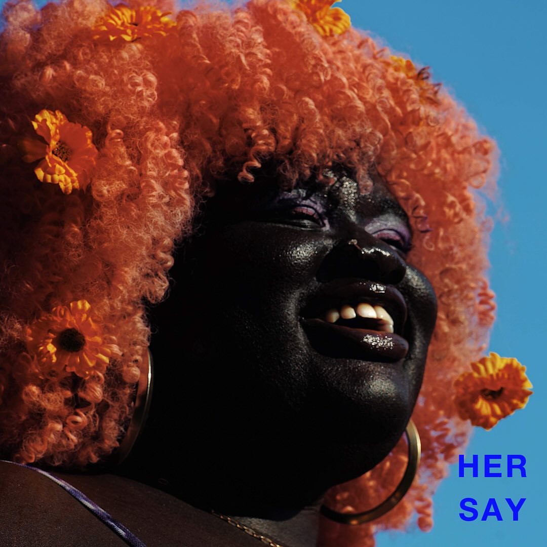 Close-up of a smiling Black woman with bright orange curly hair adorned with marigold flowers against a clear blue sky.