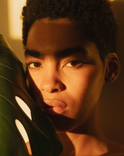 Close-up portrait of a young adult with short, dark hair, partially obscured by a large monstera leaf in warm, golden lighting.