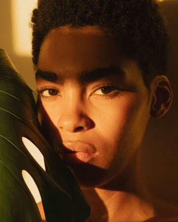 Close-up portrait of a young adult with short, dark hair, partially obscured by a large monstera leaf in warm, golden lighting.