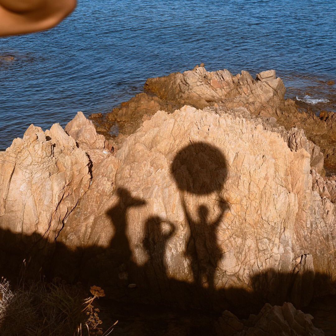 Silhouettes of three people on rocks by the sea, one holding a round object, with blue ocean water in the background.