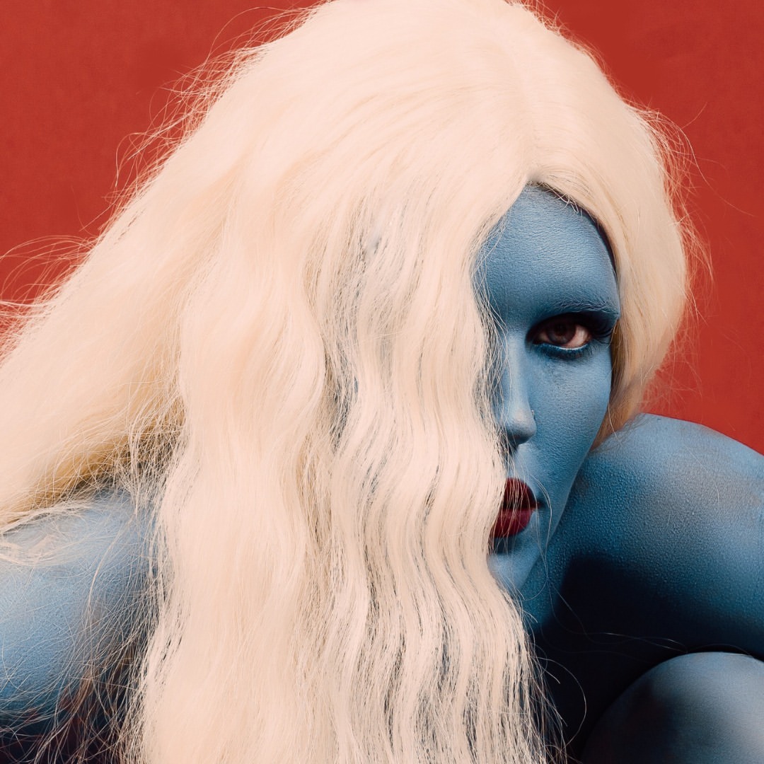 Artistic portrait of a woman with blue skin and a light blonde wig against a red background in an indoor environment.