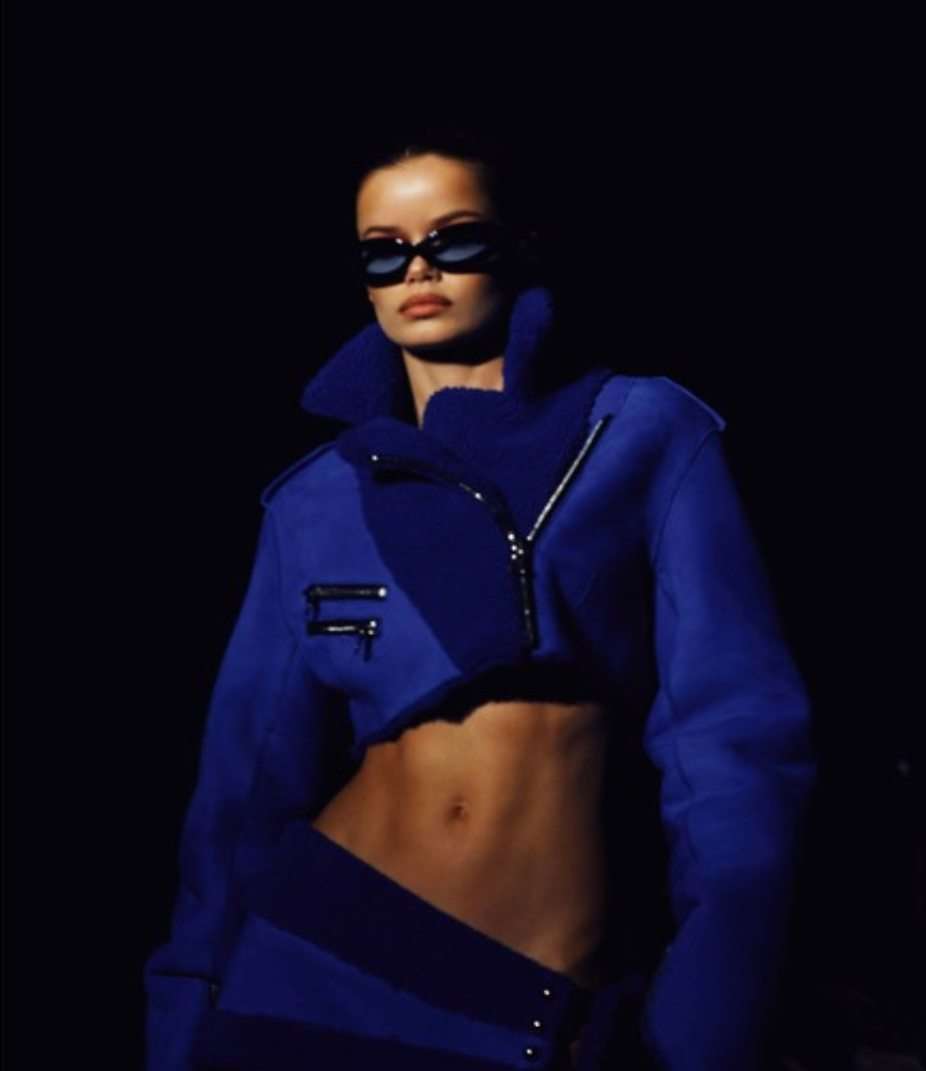 A brunette woman models a blue cropped jacket and matching bottoms with visible abs against a dark background, wearing sunglasses.