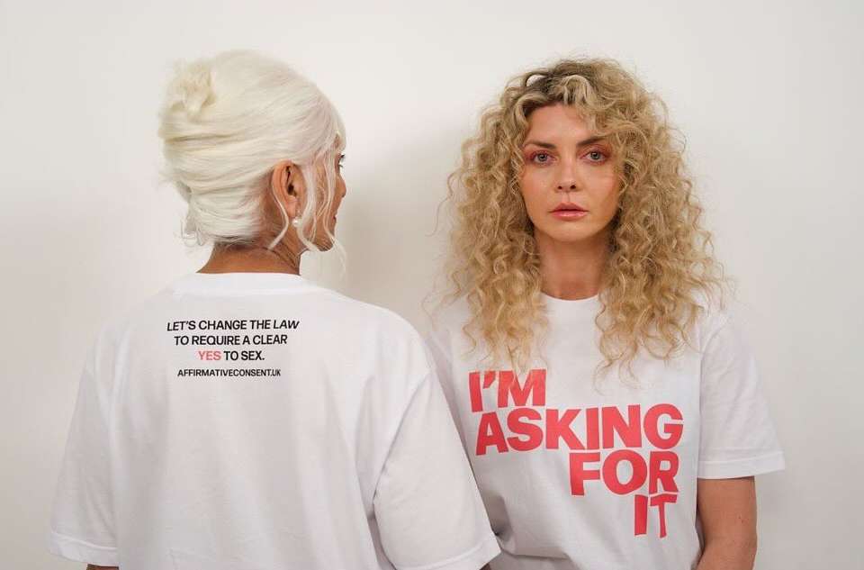 Two women stand in front of a white background, wearing white t-shirts printed with text in black and red, facing the camera.