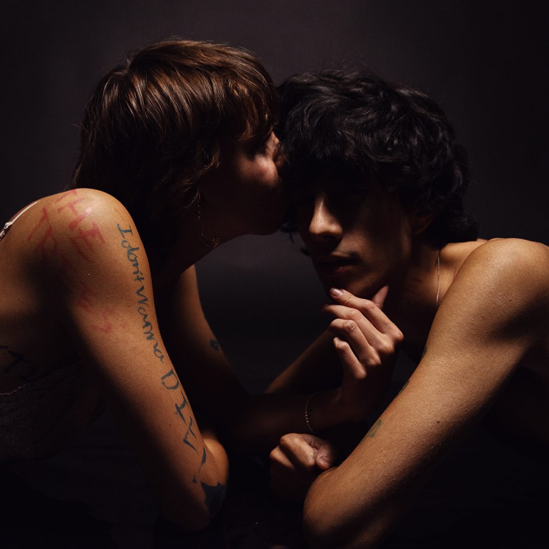 Intimate portrait of a young adult couple in a close embrace against a dark background, featuring tattoos and jewelry.