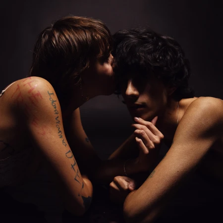 Intimate portrait of a young adult couple in a close embrace against a dark background, featuring tattoos and jewelry.